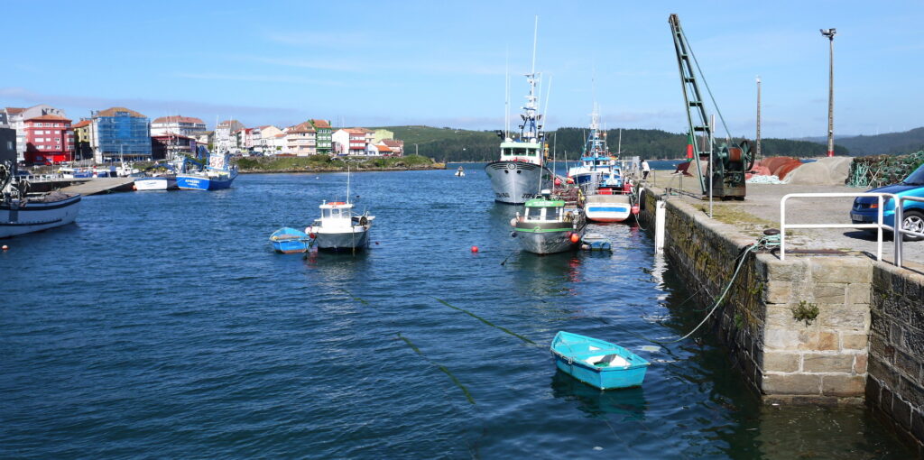Mehrere Fischerboote liegen an einem Steinpier; im Hintergrund bunte Küstenhäuser unter blauem Himmel.