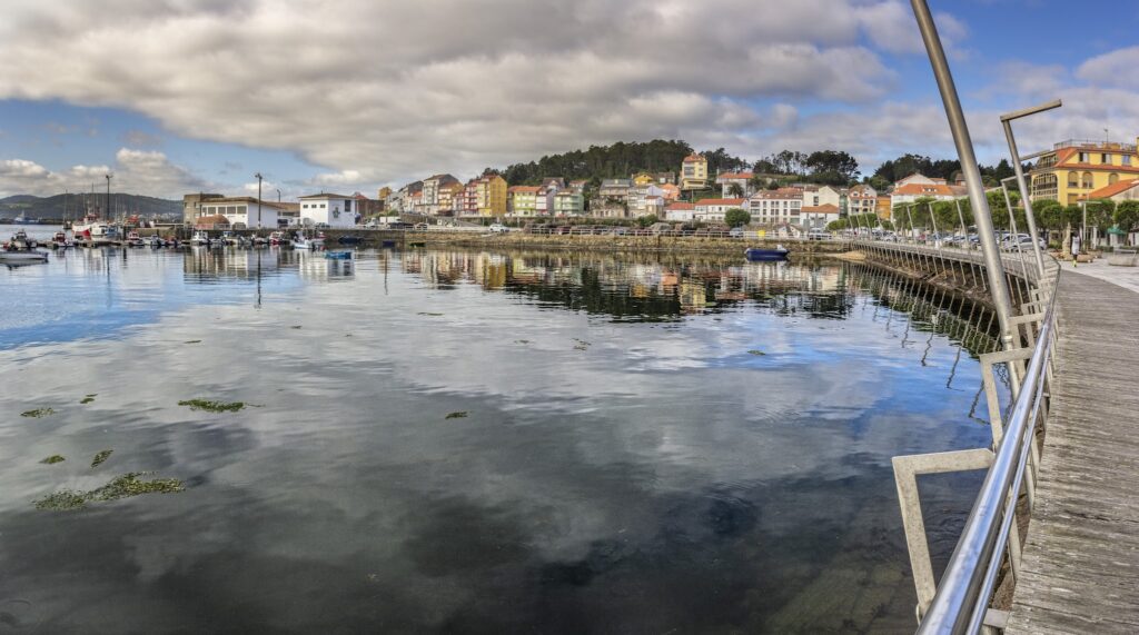 Ruhiger Hafen mit kleinen Booten, bunter Häuserreihe am Ufer und Holzpromenade, Wolken spiegeln sich im Wasser.