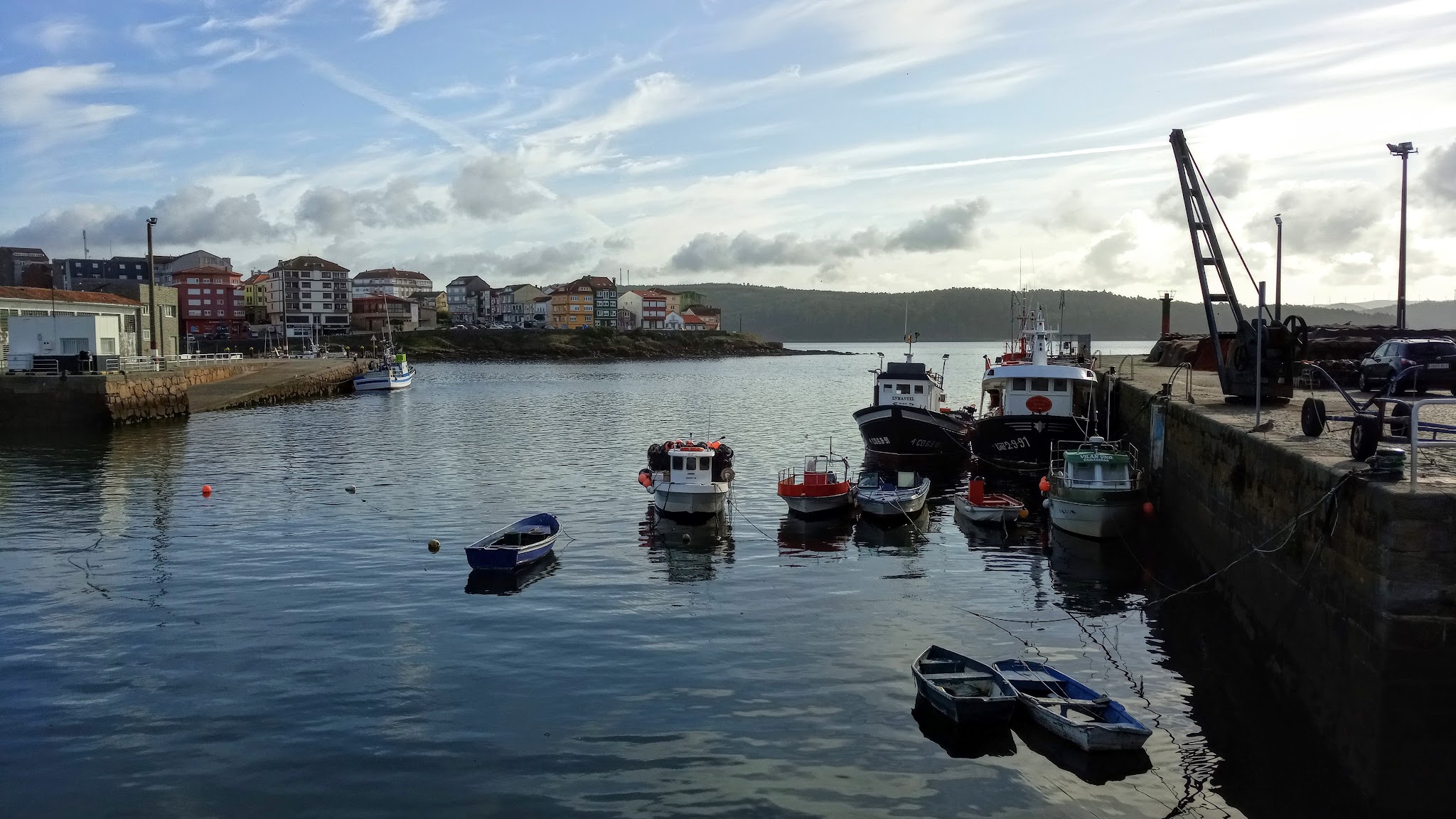 Ruhiger Hafen mit mehreren kleinen Fischerbooten am Kai, bunte Küstenhäuser und bewölkter Himmel im Hintergrund.