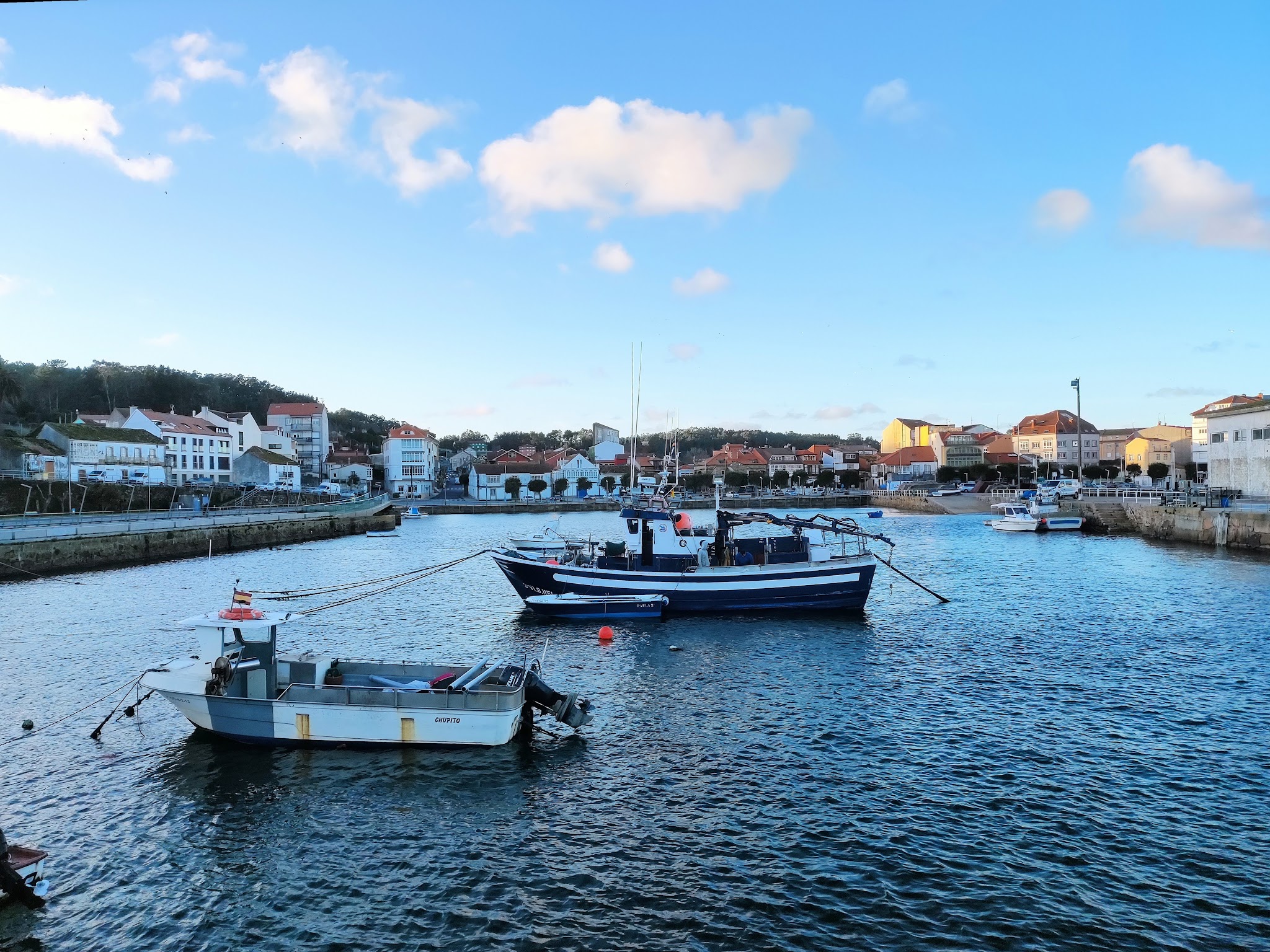 Zwei Fischerboote liegen vertäut im ruhigen Hafen, dahinter eine Küstenortschaft unter blauem Himmel.