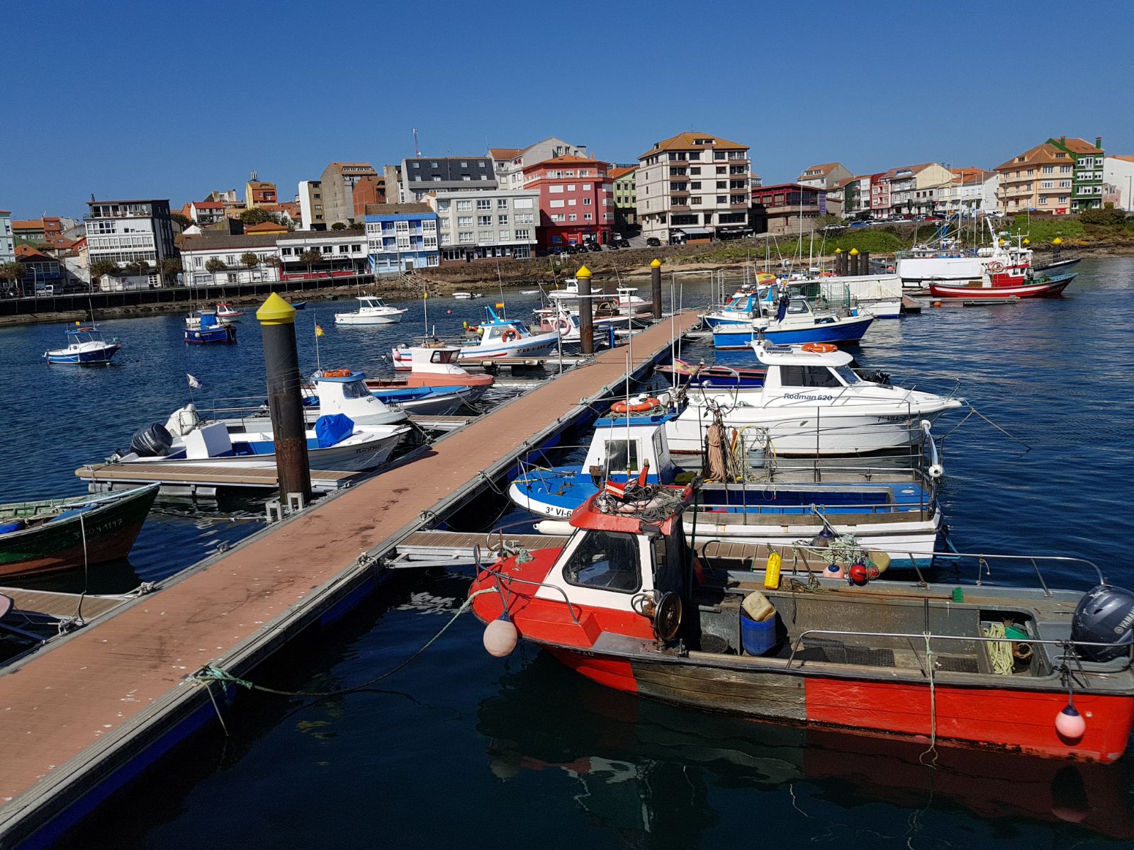 Bunte Fischer- und Sportboote am Steg eines kleinen Hafens; dahinter farbige Küstenhäuser unter klarem Himmel