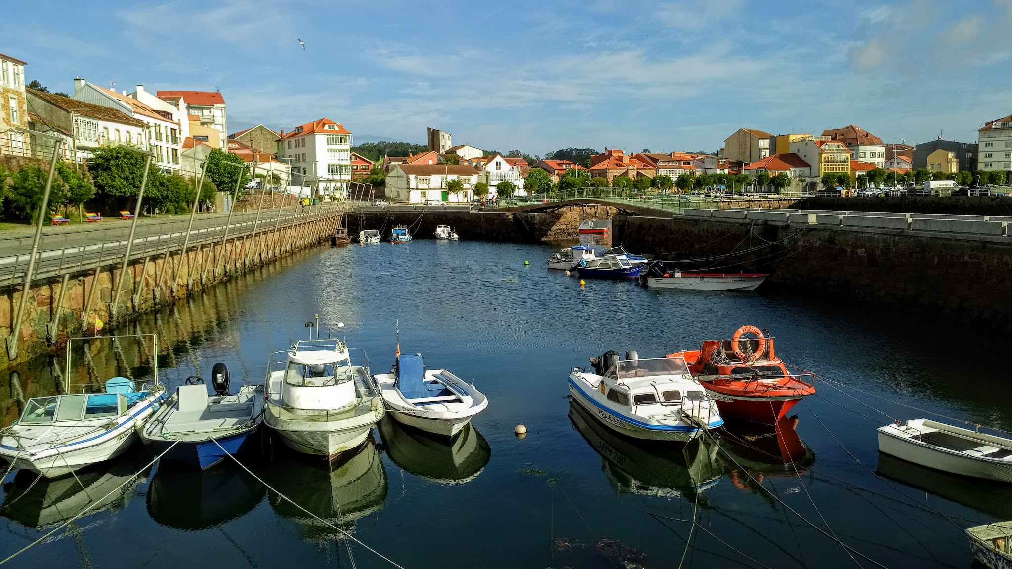 Kleiner Hafen unter blauem Himmel; mehrere festgemachte Motorboote, dahinter Uferpromenade mit bunten Häusern.