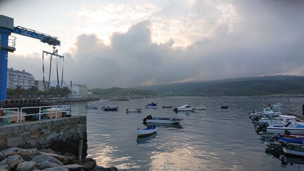 Hafen bei Dämmerung; kleine Boote dümpeln im ruhigen Wasser, links ein blauer Bootskran, Berge im Hintergrund