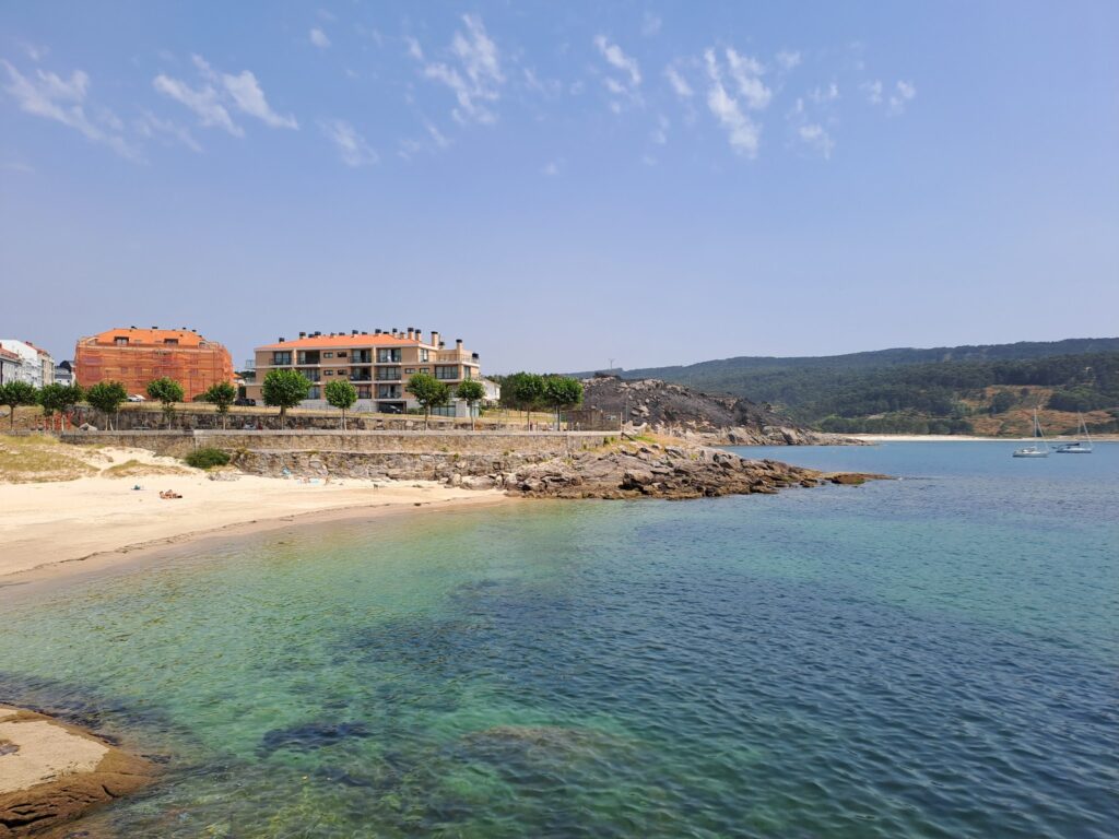 Ruhige Bucht mit türkisfarbenem Wasser, kleinem Sandstrand, Felsen, Wohnhäusern und zwei vor Anker liegenden Segelbooten.