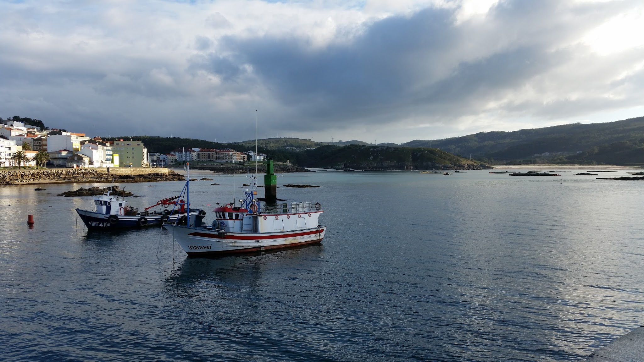 Zwei kleine Fischerboote liegen im ruhigen Hafen vor einer Küstenstadt, dahinter grüne Hügel und wolkiger Himmel.