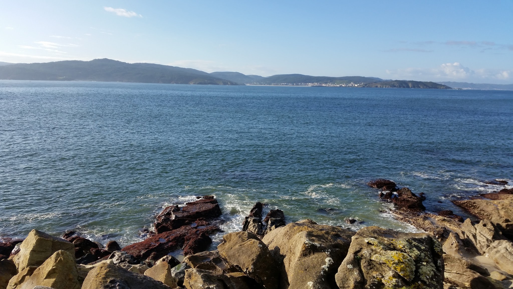 Felsige Küste, Wellen schlagen gegen braune und gelbe Steine; dahinter weitläufiges blaues Meer und entfernte Hügel.