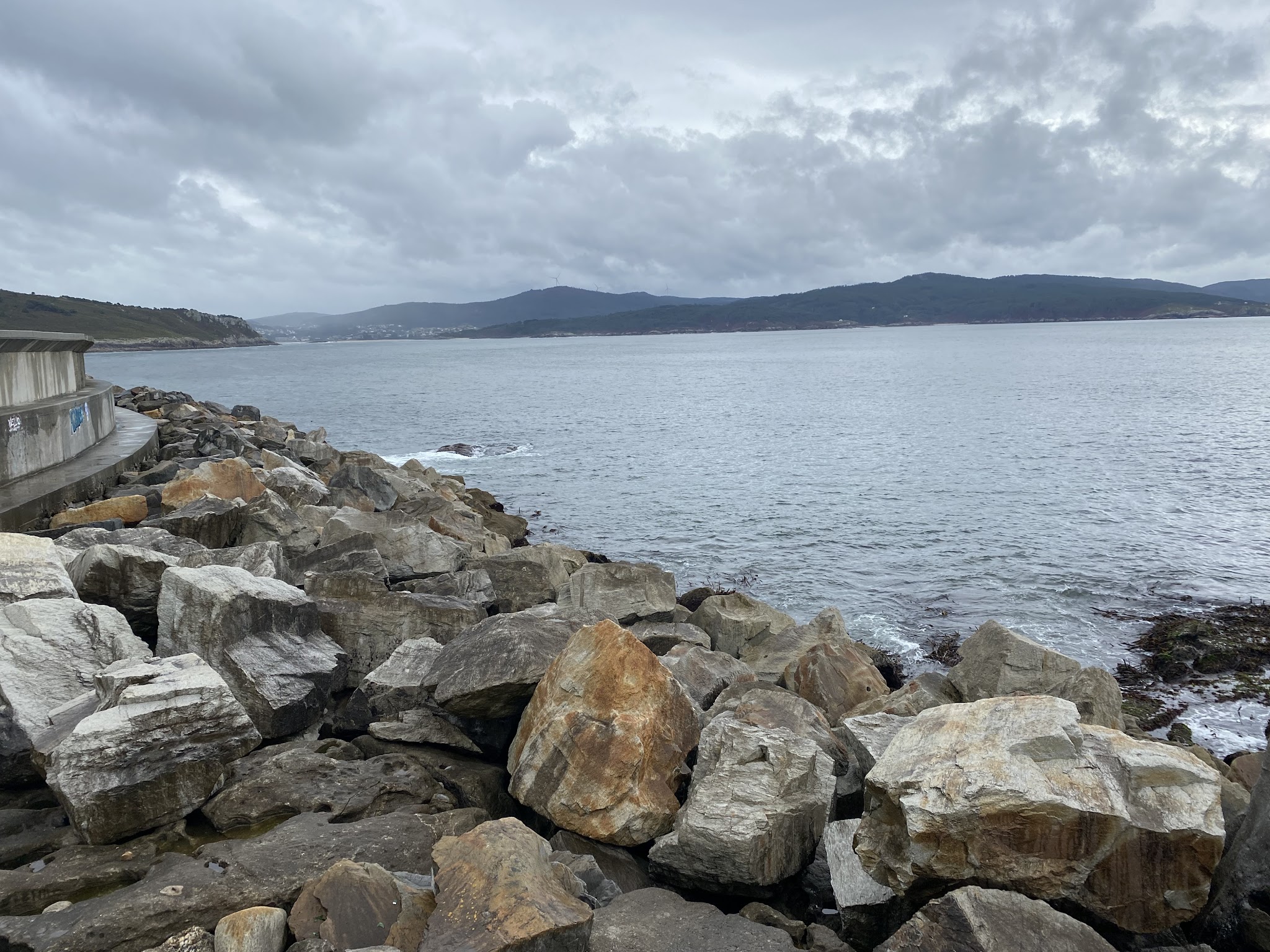 Steinige Küstenbefestigung mit großen Felsblöcken, ruhiges graues Meer unter bewölktem Himmel, Hügel am Horizont.