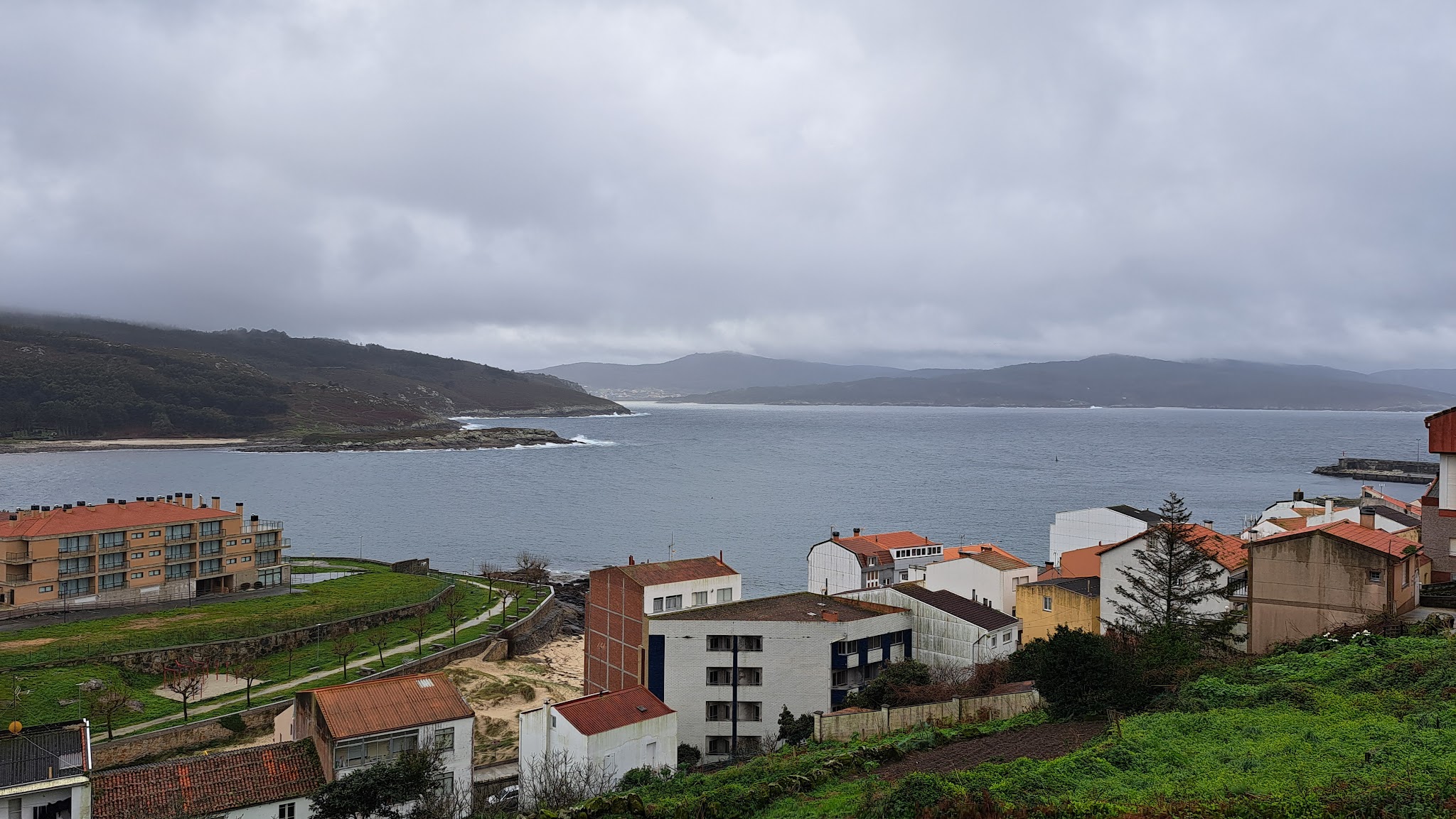 Küstendorf mit roten Ziegeldächern an grüner Böschung, graues Meer in weiter Bucht unter dicht bewölktem Himmel.