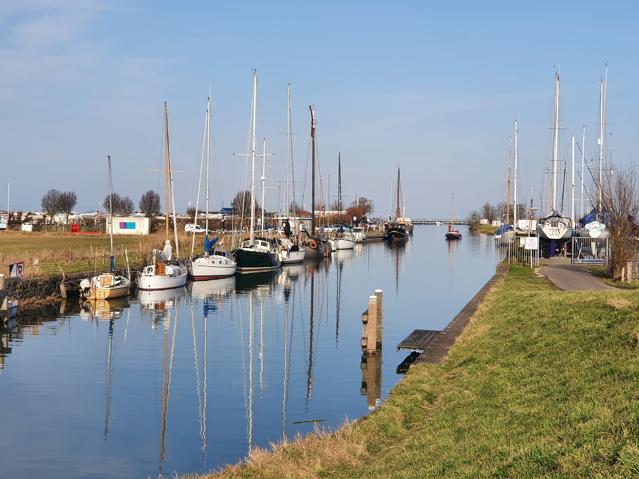 Ruhiger Kanal mit festgemachten Segelbooten; hohe Masten spiegeln sich im klaren Wasser unter blauem Himmel.