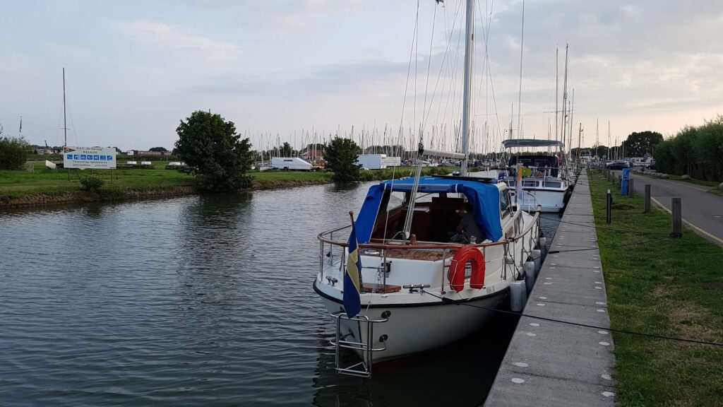 Segelboote liegen in ruhigem Kanal an Betonpier; vorn Boot mit blauer Persenning, Rettungsring, schwedischer Flagge.