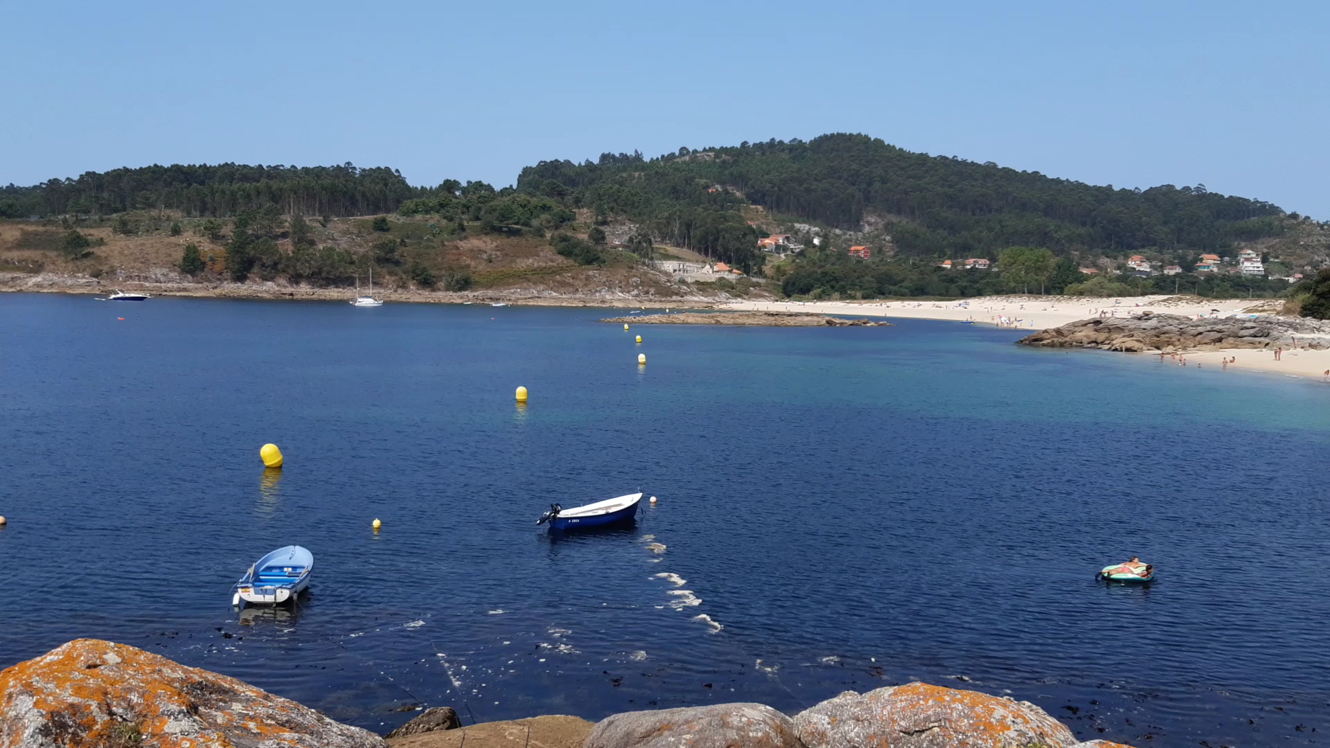 Kleine Boote und gelbe Bojen in ruhiger Bucht, dahinter Sandstrand mit Badegästen und bewaldete Hügel.
