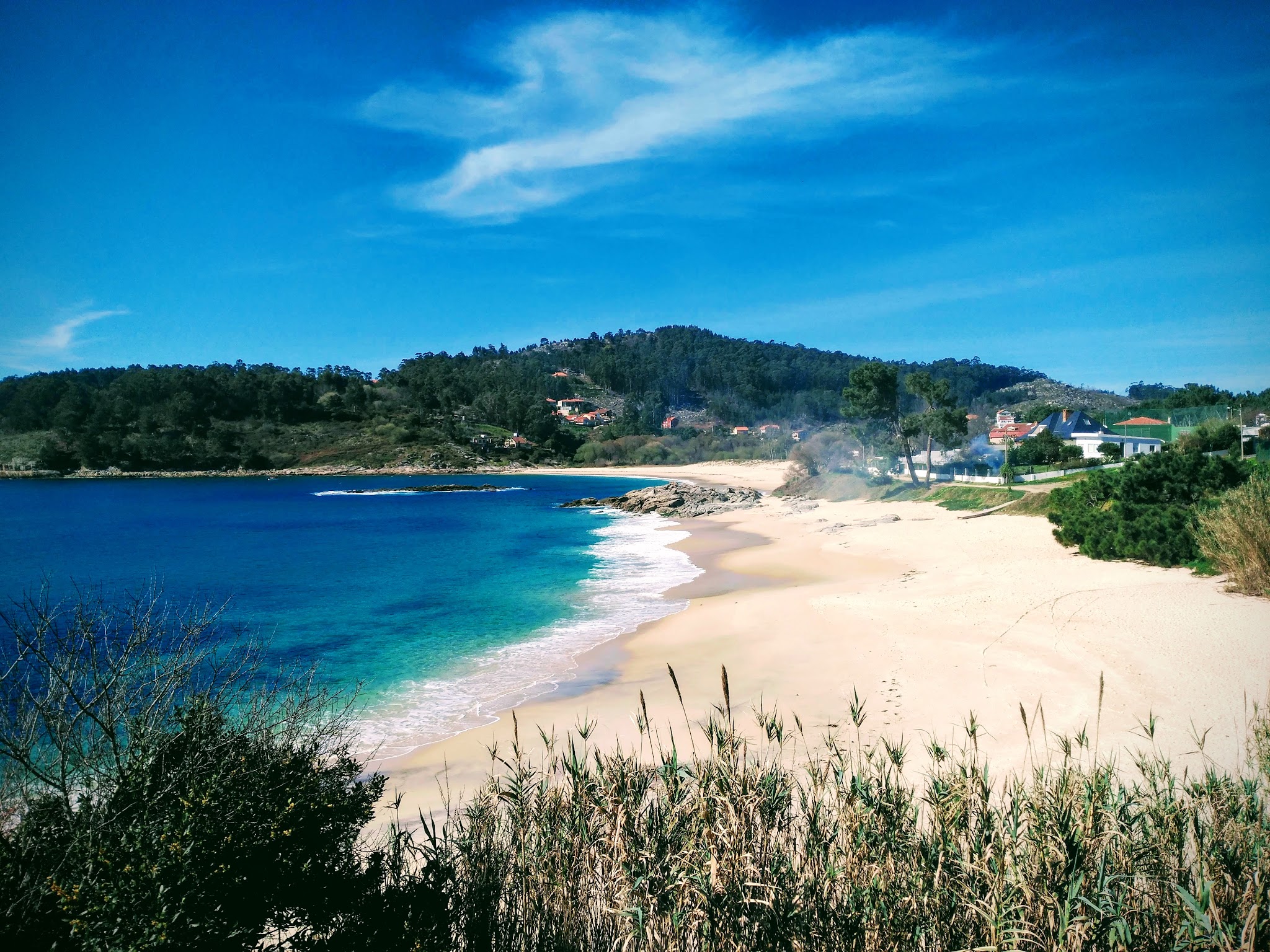 Verlassener Sandstrand mit türkisfarbenem Meer, sanften Wellen, bewaldetem Hügel und vereinzelten Häusern im Hintergrund.