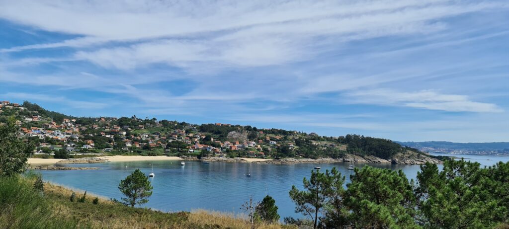 Küstenbucht mit ruhigem, blauem Wasser; Hügel voller kleiner Häuser, Sandstrand und Boote, Pinien im Vordergrund.