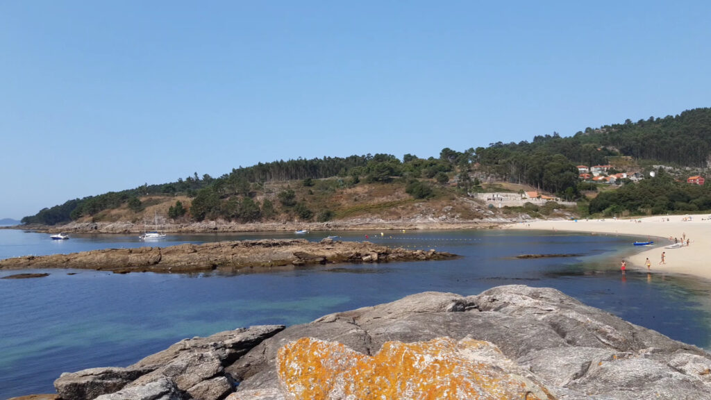 Ruhige Bucht mit klarem blauem Wasser, Felsen im Vordergrund, dahinter Sandstrand mit Badegästen und bewaldete Hügel.