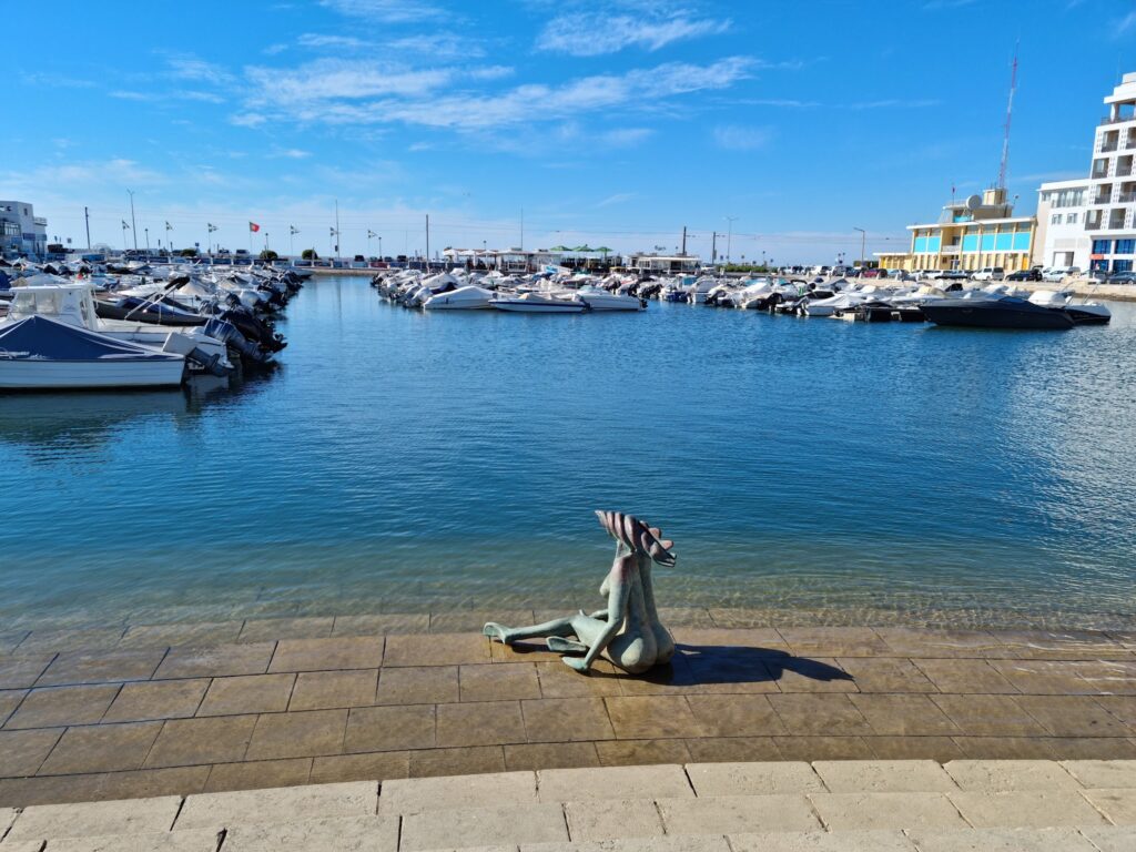 Bronzene Meerjungfrau-Skulptur sitzt am Kai eines sonnigen Jachthafens mit Reihen weißer Boote im blauen Wasser.