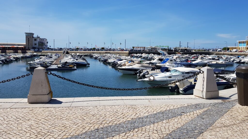 Sonnenbeschienener Yachthafen; zahlreiche Motorboote liegen ruhig an Stegen hinter einer Steinpromenade mit Ketten.