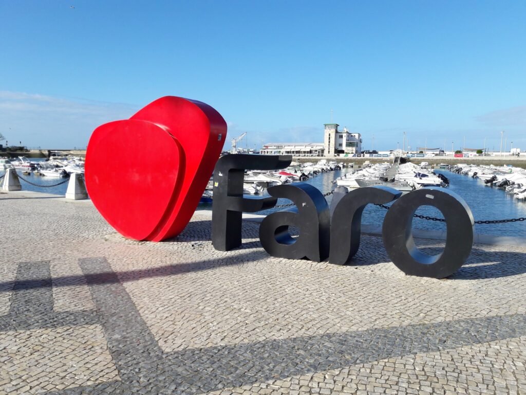 Rote Herzskulptur mit schwarzem Schriftzug „Faro“ auf Hafenpromenade; dahinter liegen Boote im sonnigen Yachthafen.