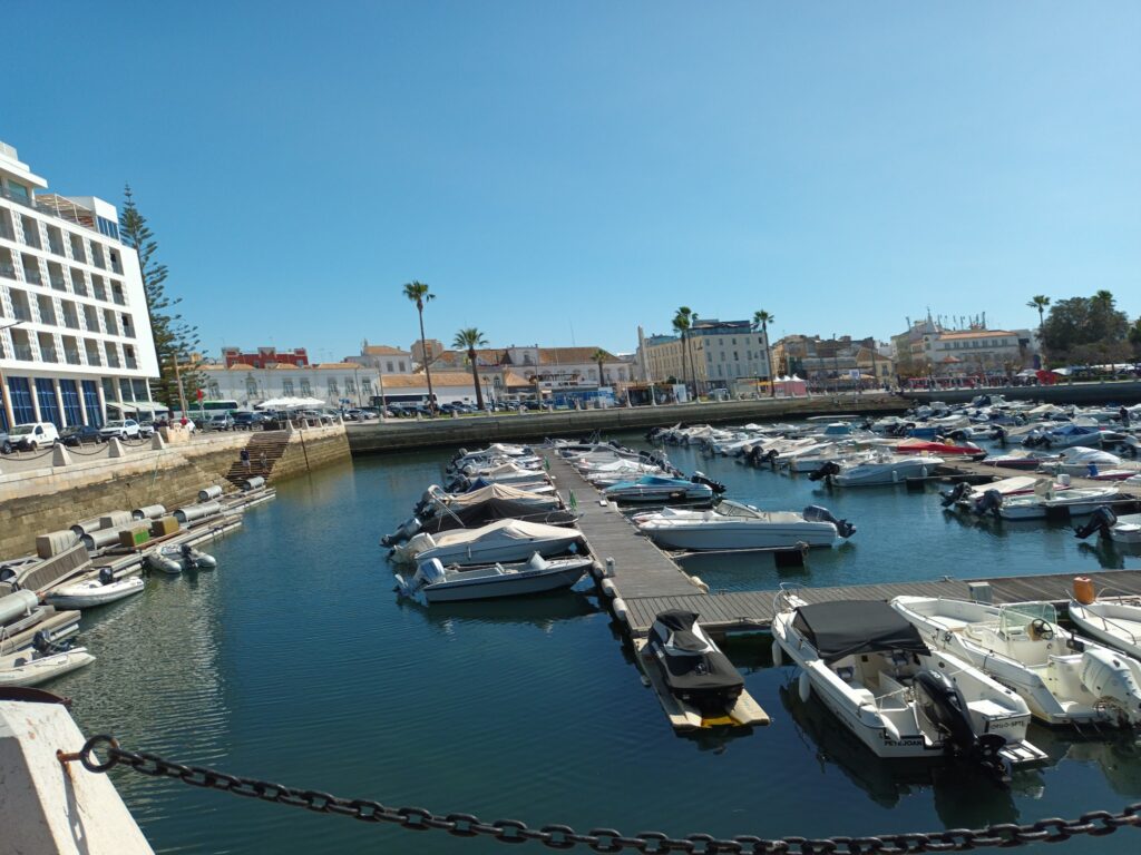 Sonnige Hafenanlage mit vielen kleinen Motorbooten an Stegen, flankiert von Uferpromenade, Hotels und Palmen.