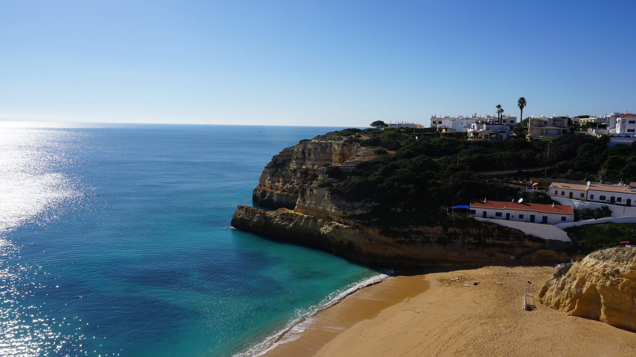 Steilklippe mit weißen Häusern über kleiner Sandbucht, türkisblaues Meer glitzert unter klarem Himmel