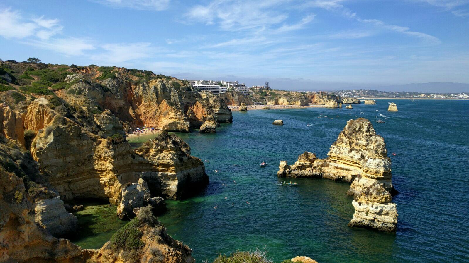 Steile, goldene Felsküste mit vorgelagerten Felsnadeln im türkisblauen Meer; Badende und Boote im Wasser.
