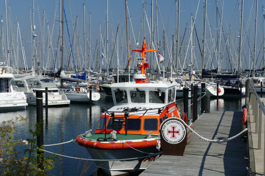 Orange-weißes Seenotrettungsboot der DGzRS liegt an Steg, im Hintergrund dichte Reihen von Segelmasten im Hafen.