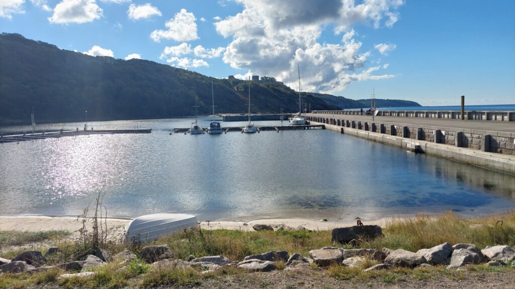 Sonnige Bucht mit langer Steinmole, vier vertäuten Segelbooten und bewaldetem Hügel im Hintergrund