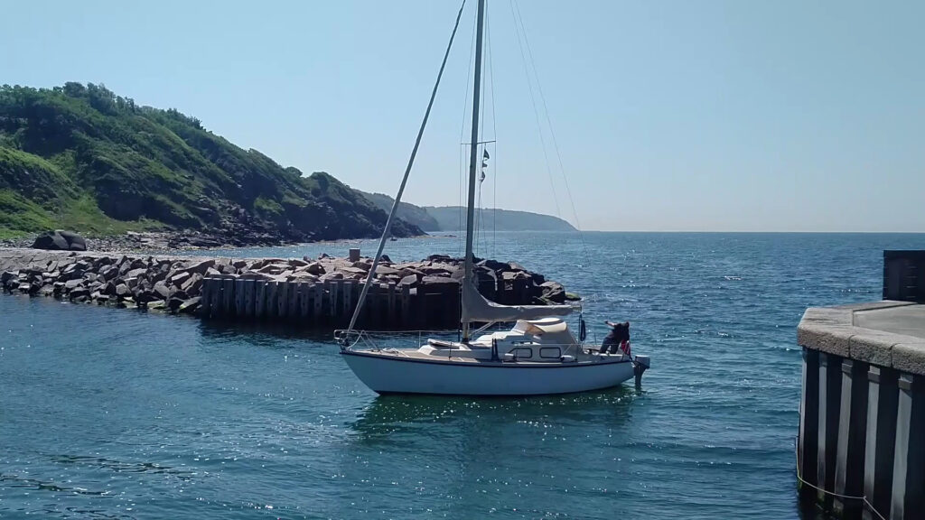 Segelboot gleitet bei Sonnenschein an felsiger Mole vorbei, grüne Klippe und blaues Meer im Hintergrund.