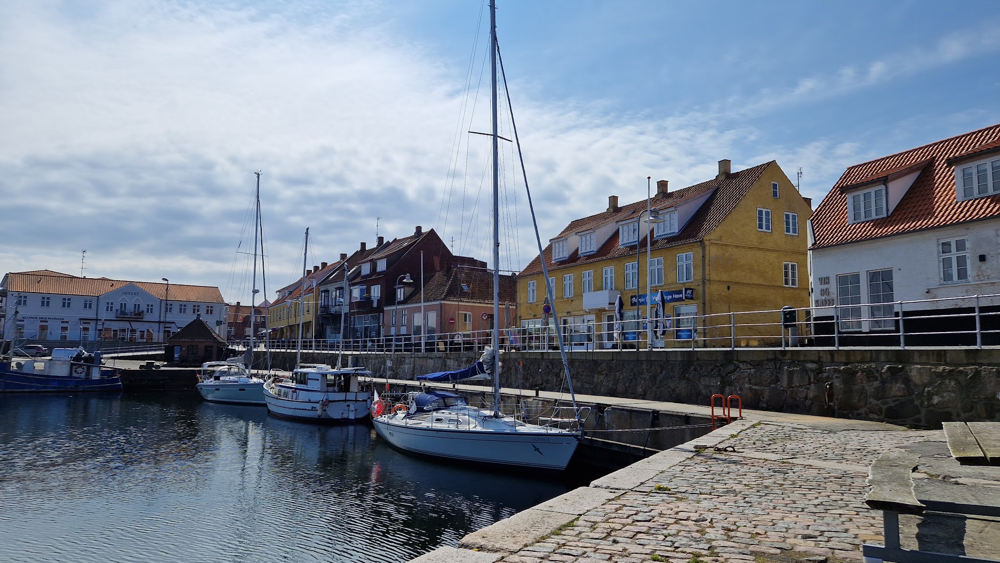 Segelboote liegen im ruhigen Hafen vor farbigen Küstenhäusern mit roten Ziegeldächern unter leicht bewölktem Himmel.
