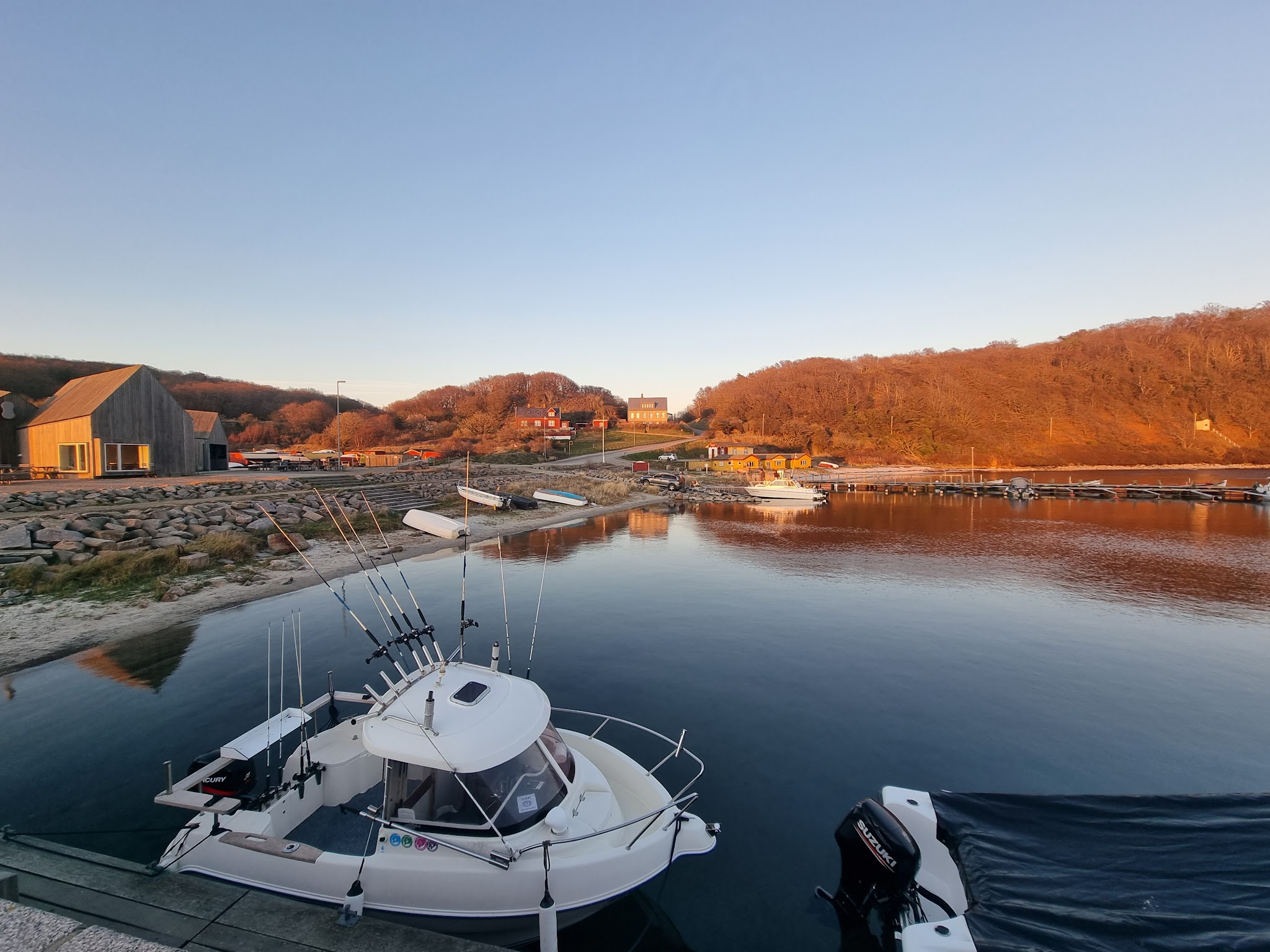 Angelboot im ruhigen Hafen; herbstliche Hänge und Holzhäuser im warmen Abendlicht spiegeln sich im Wasser.