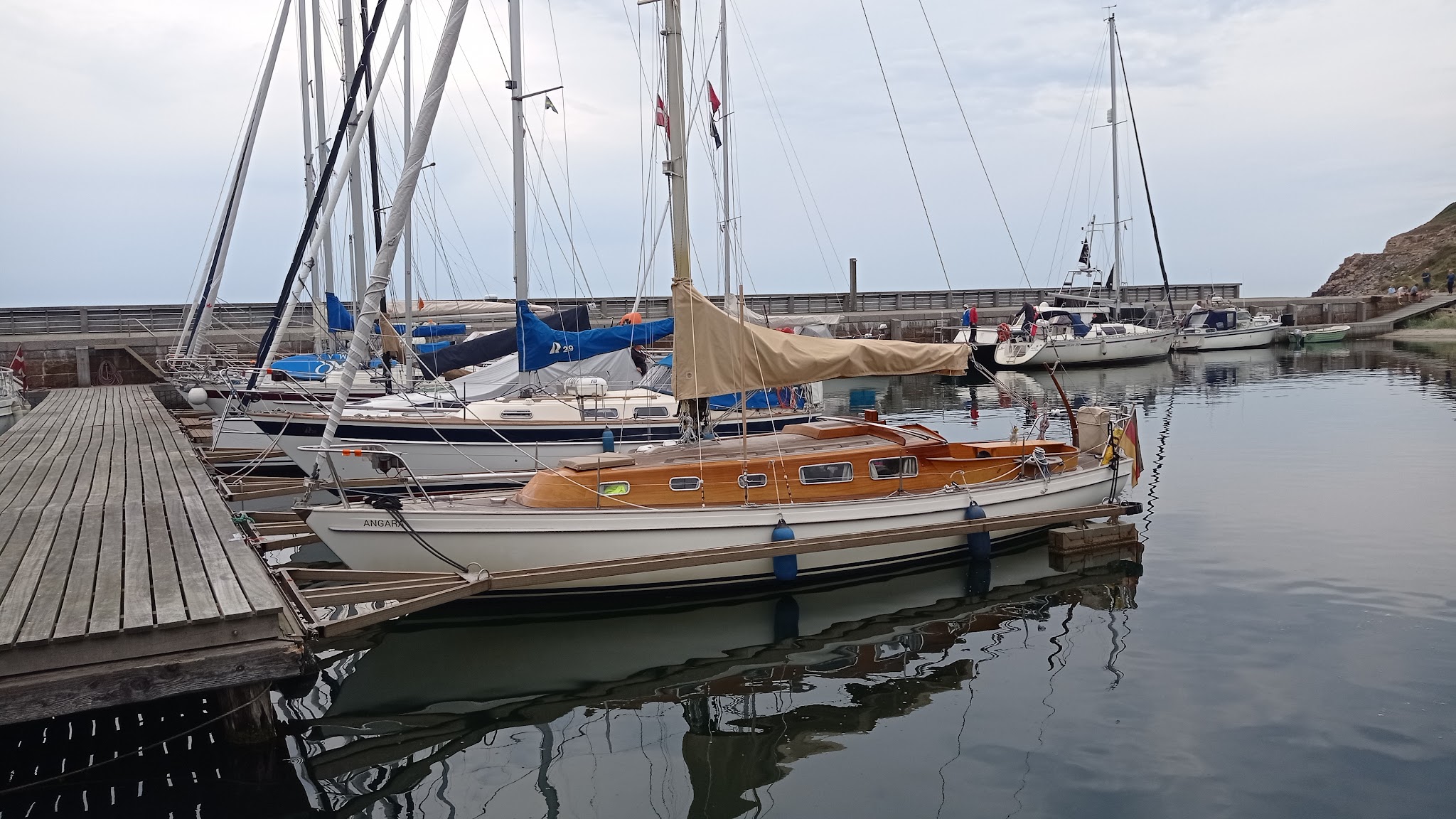 Segelboote liegen an hölzernem Steg im ruhigen Hafen, Masten spiegeln sich im stillen Wasser.