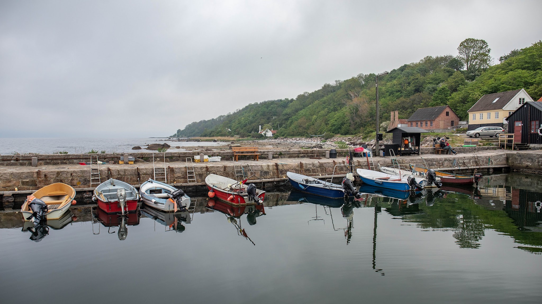 Mehrere kleine Motorboote spiegeln sich im ruhigen Hafenwasser; dahinter Küste mit grünen Hügeln und Häusern.