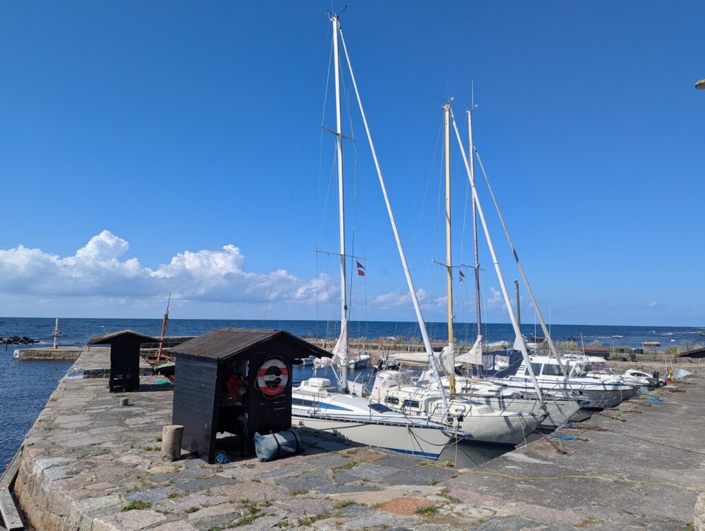 Segelboote am steinigen Kai unter blauem Himmel; zwei schwarze Holzhütten und ruhige Ostsee im Hintergrund.