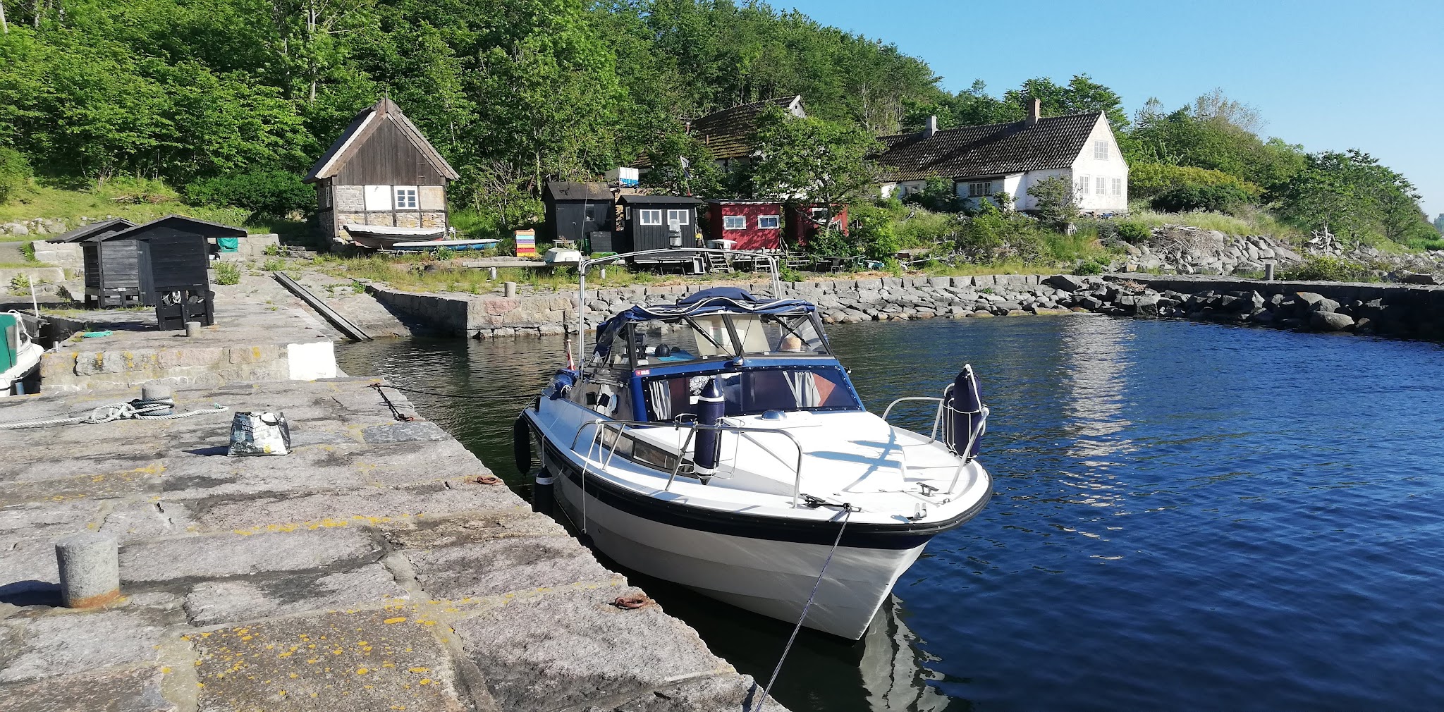 Kleine Motorjacht am steinernen Kai; dahinter Holzhütten, weißes Haus und grüner Wald im Sonnenschein.