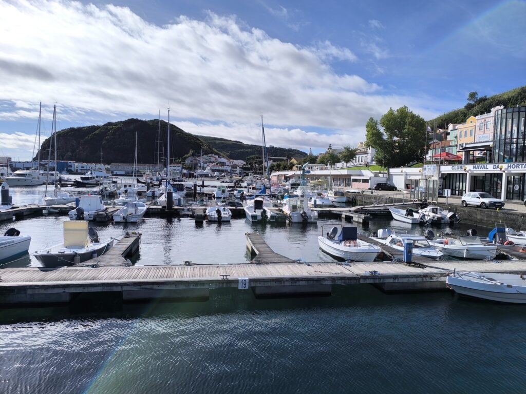 Kleiner Hafen mit anliegenden Motor- und Segelbooten, dahinter Hügel und bunte Häuser unter blauem Himmel.