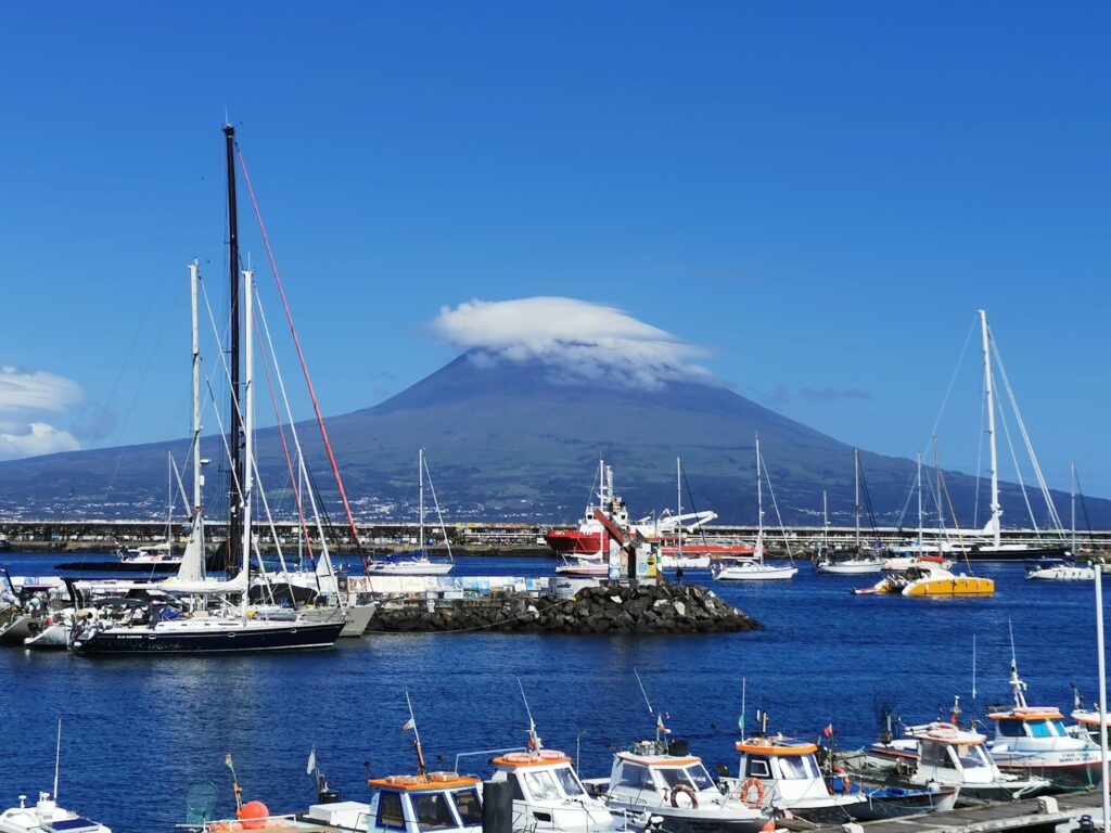 Segelboote im Hafen vor einem kegelförmigen Vulkan, dessen Gipfel von einer weißen Wolkenhaube ummantelt ist.