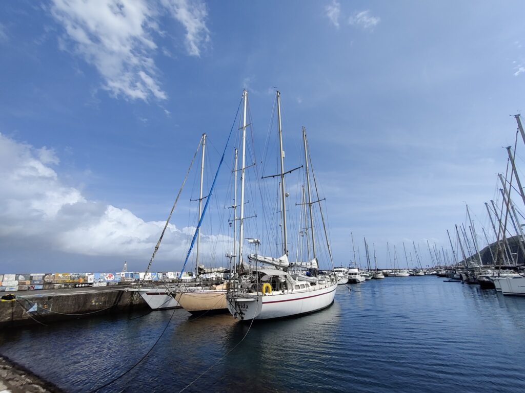 Segelboote mit hohen Masten liegen an Pier in ruhigem, sonnigen Hafen; blauer Himmel mit wenigen Wolken.