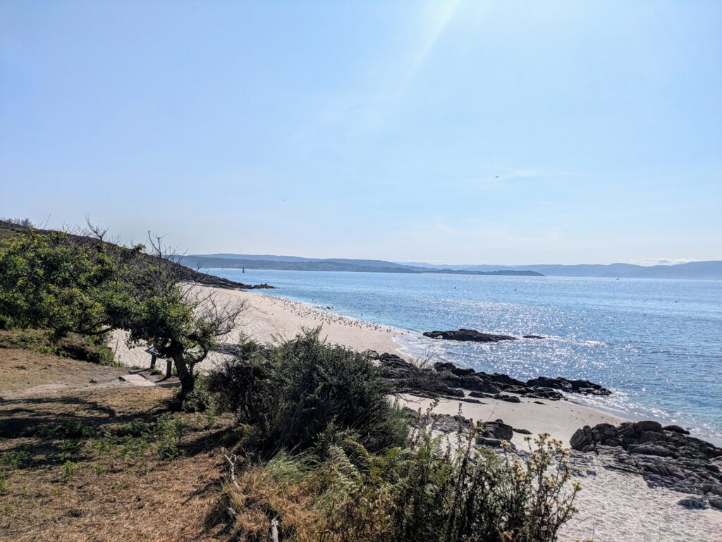 Sonniger Strand mit weißem Sand, Felsgruppen und funkelnd blauem Meer; Dünenbewuchs im Vordergrund