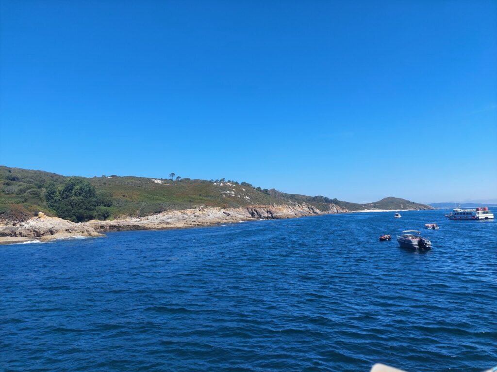 Klares Meer mit wenigen Booten vor grüner Felsküste unter wolkenlosem, strahlend blauem Himmel.