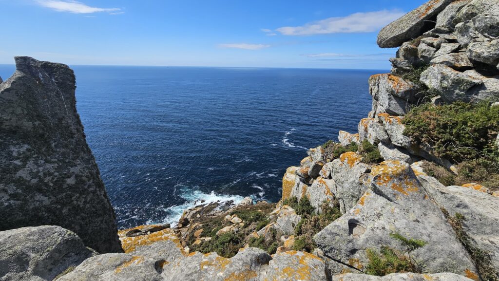 Flechten­bedeckte Klippen fallen steil zum tiefblauen Atlantik, unten Gischt, oben weiter Himmel mit einzelnen Wolken.