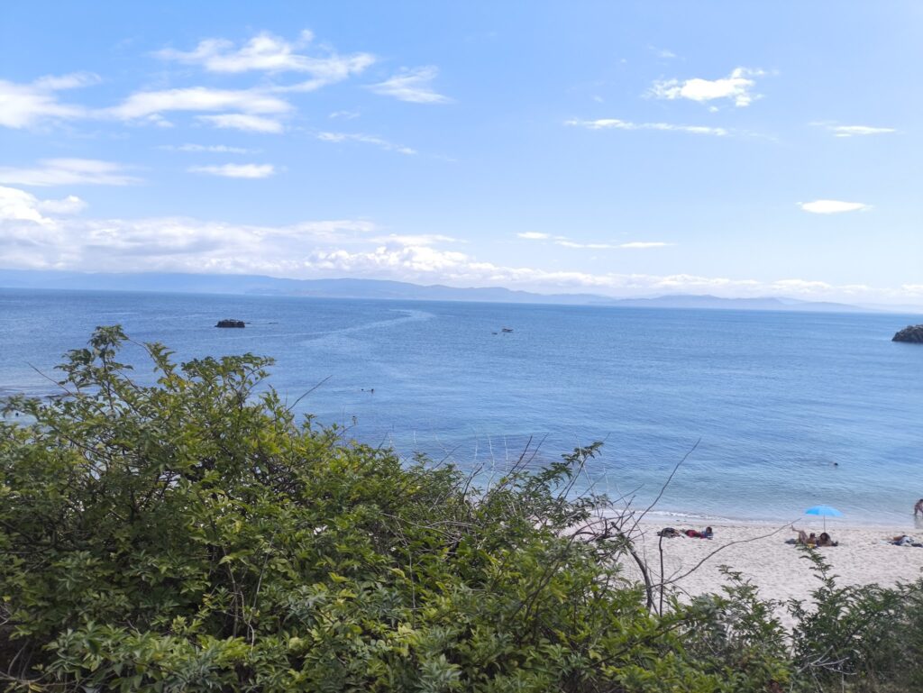 Grüne Büsche vor sandigem Strand; dahinter ruhiges blaues Meer, vereinzelte Badegäste, ferne Berge am Horizont.