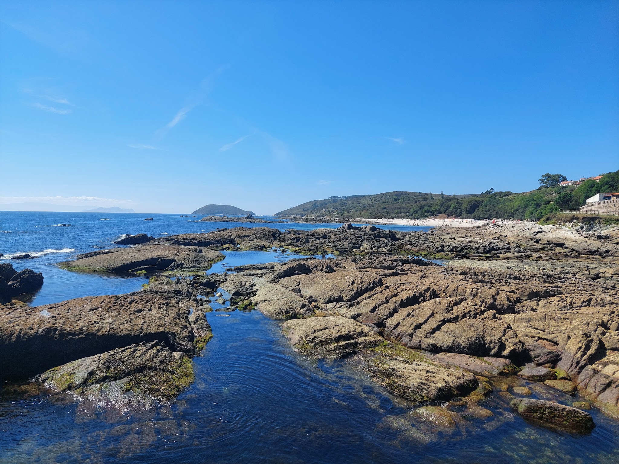 Felsige Küste mit Gezeitentümpeln vor blauem Meer; im Hintergrund Sandstrand, grüne Hügel und klarer Himmel.