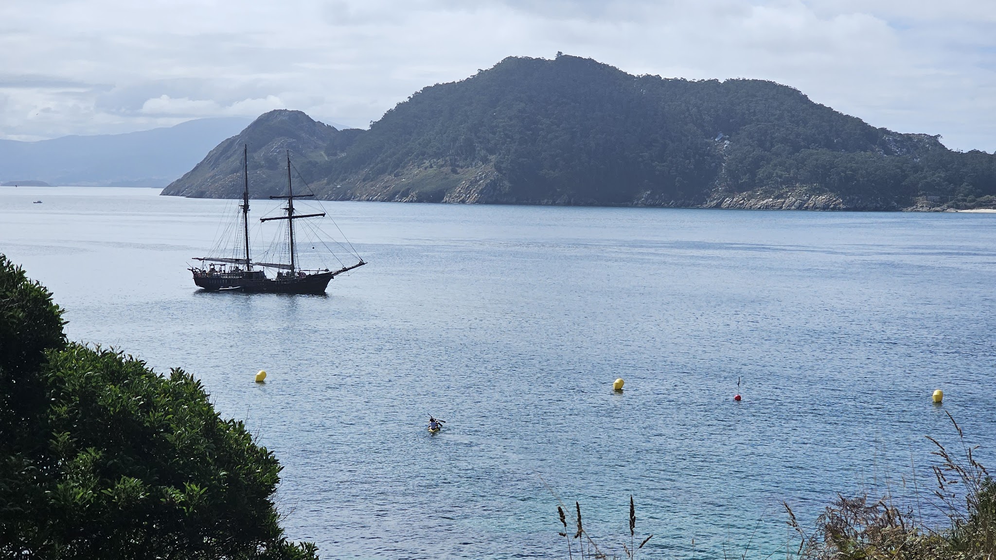 Zweimastiges Segelschiff liegt in ruhiger Bucht vor bewaldeter Insel; Kajakfahrer und gelbe Bojen im klaren Wasser.