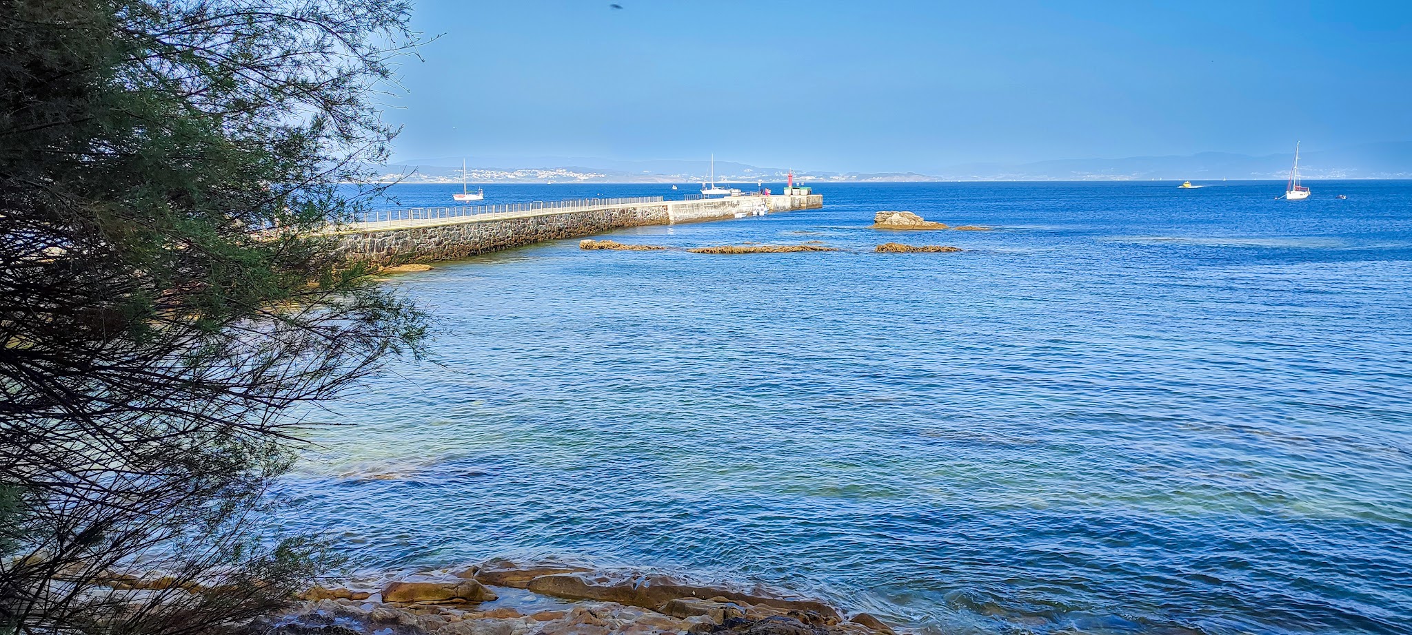 Blaues Meer mit Steinpier und kleinem Leuchtturm, vereinzelt segelnde Boote; Felsküste und Baumzweige im Vordergrund.