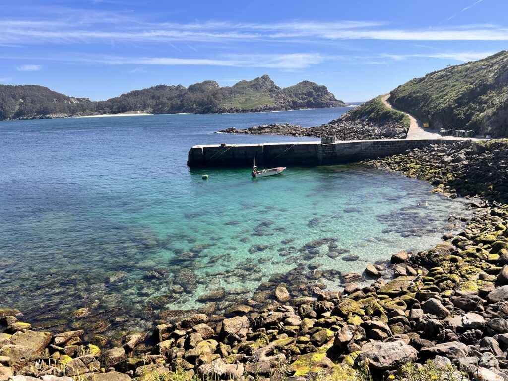 Türkisfarbene Bucht mit kleinem Boot an Steinpier, von Felsen und grünen Hügeln umgeben, blauer Himmel