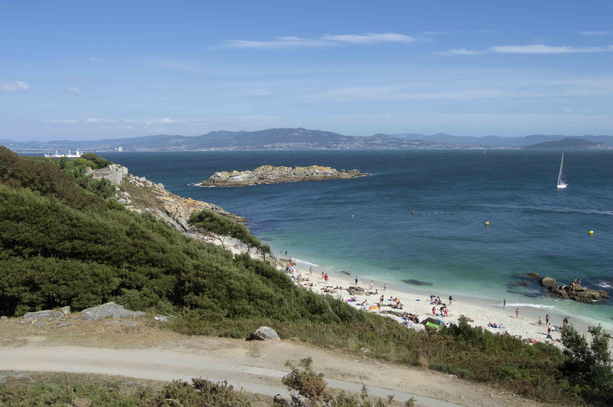 Badegäste an weißem Sandstrand, türkisblaues Meer mit Segelboot, vorgelagerter Felsen, Berge am Horizont.