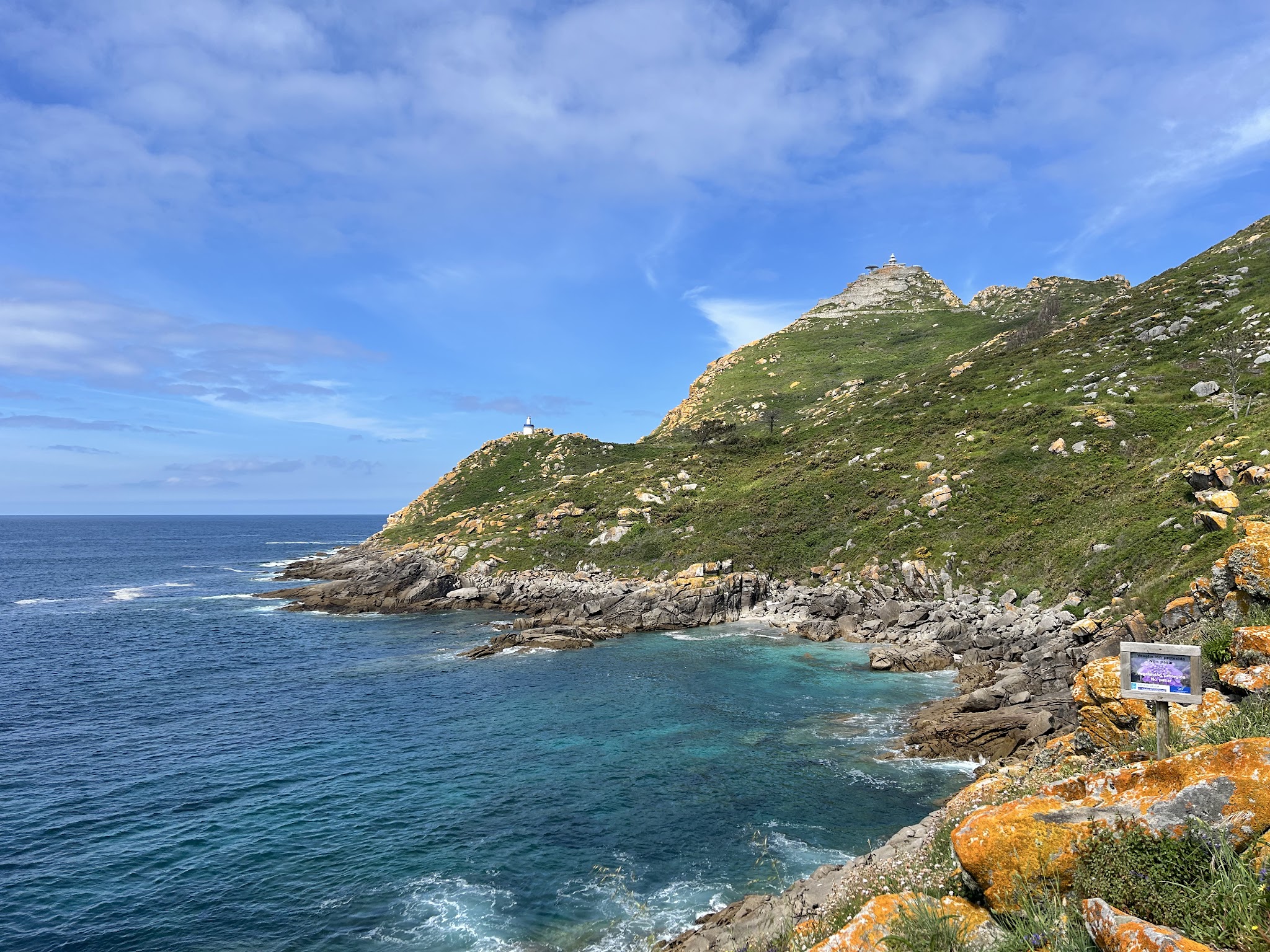 Klippenküste mit türkisblauem Meer, grüner Felsenhügel; winziger Leuchtturm auf der Spitze unter blauem Himmel.