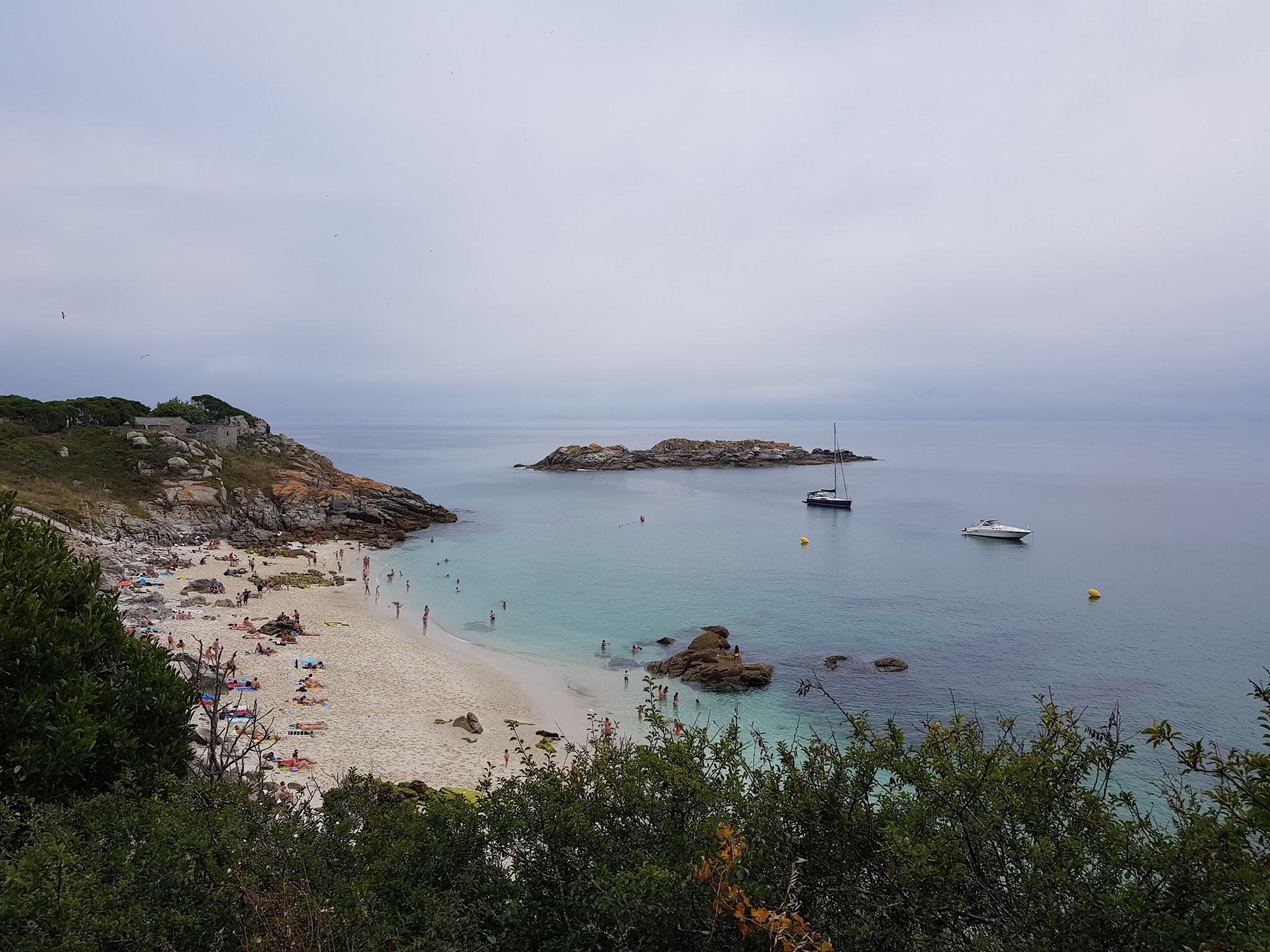 Kleine Bucht mit hellem Sand, Badenden im türkisblauen Wasser, vorgelagertem Felsen und zwei ankernden Booten.