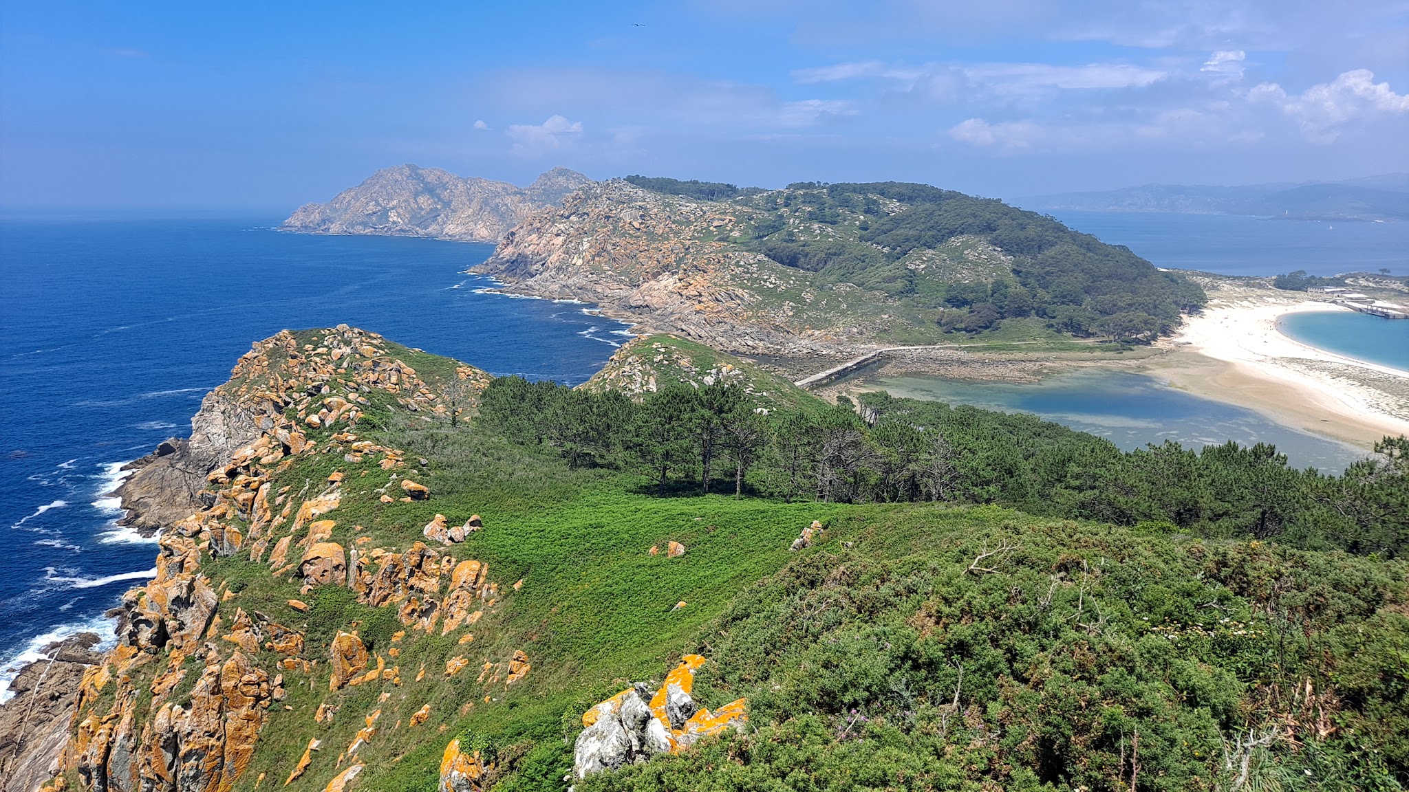Grüne Klippeninsel mit zerklüfteten Felsen, Pinien, türkisblauem Meer und geschwungener Sandbucht bei klarem Himmel.