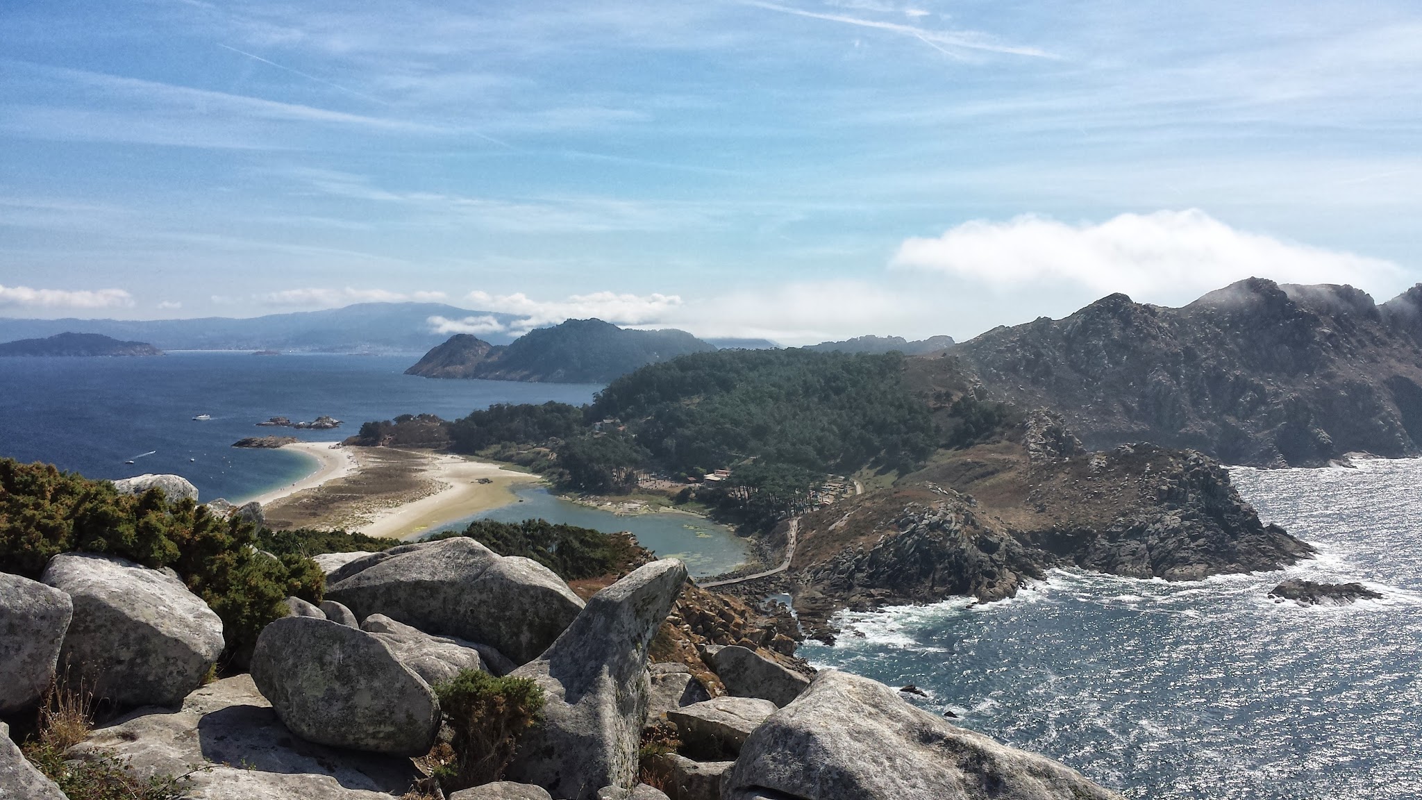 Felsige Küste mit vorgelagertem Sandstreifen, grünem Inselhügel, glitzerndem Meer und fernen Bergen unter blauem Himmel