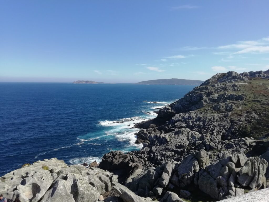 Felsküste mit schroffen Klippen, darunter tiefblaues Meer mit weißen Wellen, im Hintergrund ferne Inseln und blauer Himmel